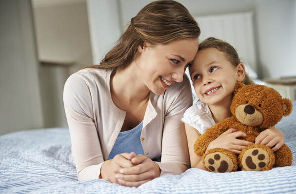nanny with a little girl holding a teddy bear lying on a bed looking at each other and smiling